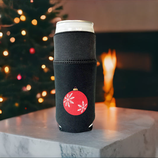 All black Candabra drink sleeve with a red ornament and white snowflake design.  Pictured on a slim 12 oz can with the elastic cuff extended in the background is a Christmas tree and cozy fire.